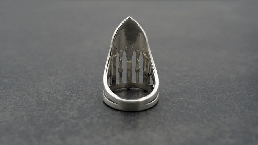 A clean studio shot of a tall, architectural sterling silver ring standing on a matte grey surface. The ring creates the silhouette of a Gothic "Lancet Window." It features three vertical openings separated by slender bars (mullions), topped by an intricate geometric tracery design within the pointed arch. The metal is matte silver with dark oxidation in the corners. (etsy image id: 7500328125)