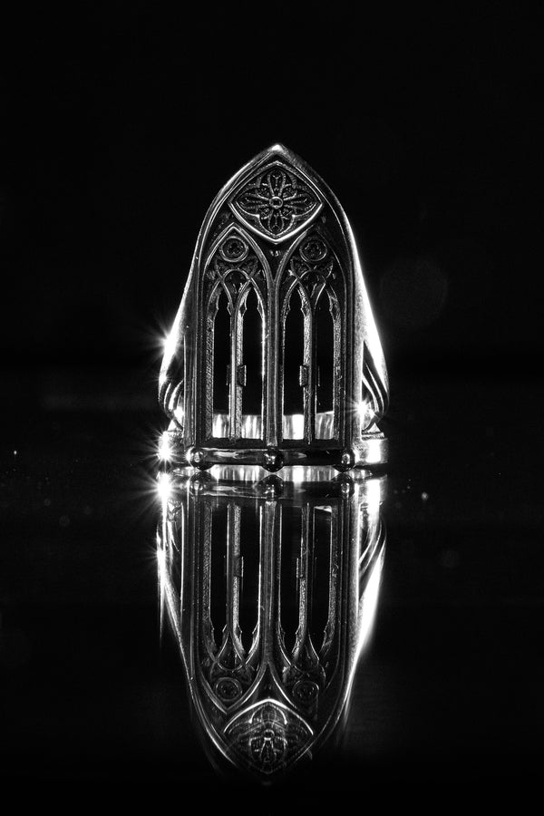 A clean studio shot of a tall, architectural sterling silver ring standing on a matte grey surface. The ring creates the silhouette of a Gothic "Lancet Window." It features three vertical openings separated by slender bars (mullions), topped by an intricate geometric tracery design within the pointed arch. The metal is matte silver with dark oxidation in the corners. (etsy image id: 7500328125)