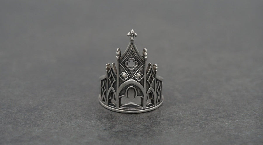 A clean studio shot of a sculptural sterling silver ring resting on a matte grey surface. The ring depicts a Gothic building facade with a tall, triangular central peak (gable) filled with geometric cross-shaped tracery. Flanking the center are lower pointed arches and vertical buttresses that wrap around the band. The silver is matte-finished with dark oxidation in the recesses to mimic stone shadows. (etsy image id: 7452370200)