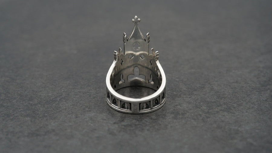 A clean studio shot of a sculptural sterling silver ring resting on a matte grey surface. The ring depicts a Gothic building facade with a tall, triangular central peak (gable) filled with geometric cross-shaped tracery. Flanking the center are lower pointed arches and vertical buttresses that wrap around the band. The silver is matte-finished with dark oxidation in the recesses to mimic stone shadows. (etsy image id: 7452370200)