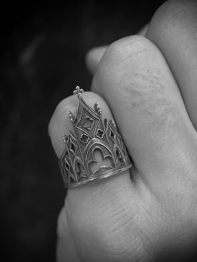 A clean studio shot of a sculptural sterling silver ring resting on a matte grey surface. The ring depicts a Gothic building facade with a tall, triangular central peak (gable) filled with geometric cross-shaped tracery. Flanking the center are lower pointed arches and vertical buttresses that wrap around the band. The silver is matte-finished with dark oxidation in the recesses to mimic stone shadows. (etsy image id: 7452370200)