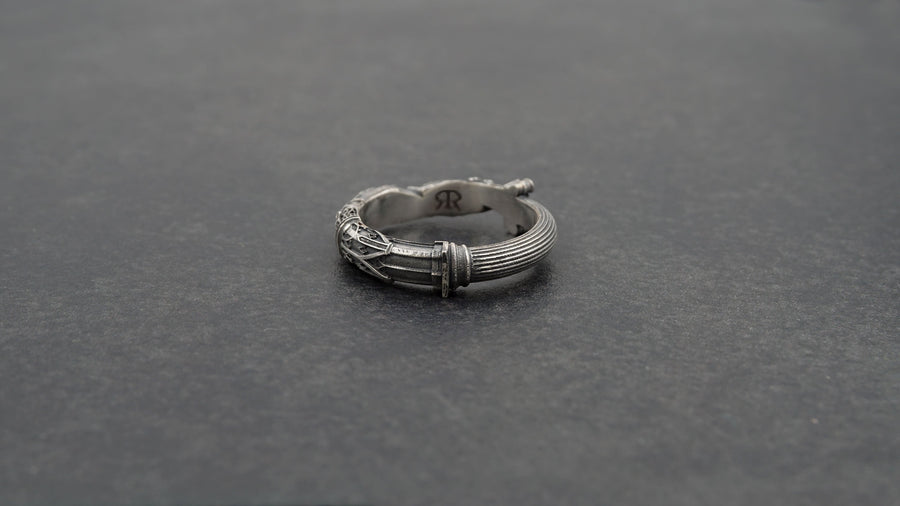 A cinematic studio shot of a sculptural sterling silver ring resting on a dark textured fabric. The focal point is two realistic hands clasped in a handshake (the Fede motif). Instead of fabric sleeves, the wrists emerge from architectural column capitals with distinct "volute" scrolls (Ionic style). The band continues as a textured, fluted column shaft. The silver is heavily oxidized to mimic ancient stone. (etsy image id: 7452415972)