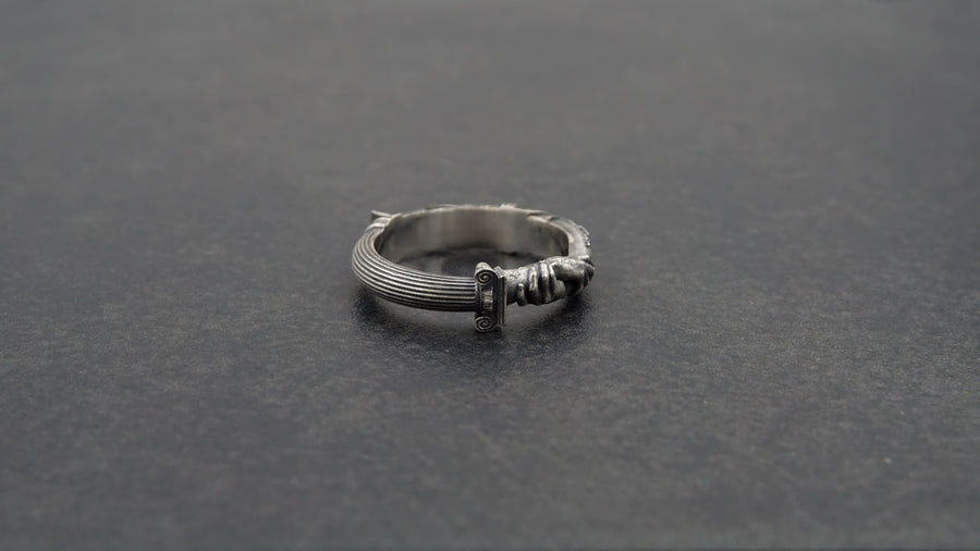 A cinematic studio shot of a sculptural sterling silver ring resting on a dark textured fabric. The focal point is two realistic hands clasped in a handshake (the Fede motif). Instead of fabric sleeves, the wrists emerge from architectural column capitals with distinct "volute" scrolls (Ionic style). The band continues as a textured, fluted column shaft. The silver is heavily oxidized to mimic ancient stone. (etsy image id: 7452415972)