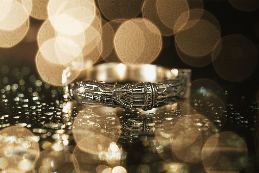 A cinematic studio shot of a sculptural sterling silver ring resting on a dark textured fabric. The focal point is two realistic hands clasped in a handshake (the Fede motif). Instead of fabric sleeves, the wrists emerge from architectural column capitals with distinct "volute" scrolls (Ionic style). The band continues as a textured, fluted column shaft. The silver is heavily oxidized to mimic ancient stone. (etsy image id: 7452415972)