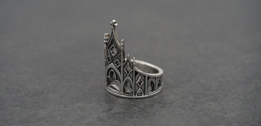 A clean studio shot of a sculptural sterling silver ring resting on a matte grey surface. The ring depicts a Gothic building facade with a tall, triangular central peak (gable) filled with geometric cross-shaped tracery. Flanking the center are lower pointed arches and vertical buttresses that wrap around the band. The silver is matte-finished with dark oxidation in the recesses to mimic stone shadows. (etsy image id: 7452370200)