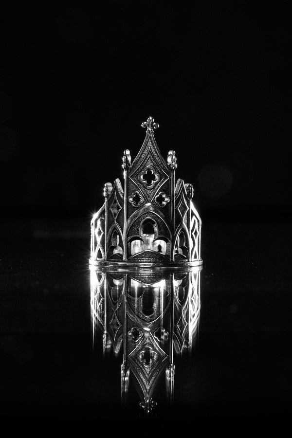 A clean studio shot of a sculptural sterling silver ring resting on a matte grey surface. The ring depicts a Gothic building facade with a tall, triangular central peak (gable) filled with geometric cross-shaped tracery. Flanking the center are lower pointed arches and vertical buttresses that wrap around the band. The silver is matte-finished with dark oxidation in the recesses to mimic stone shadows. (etsy image id: 7452370200)