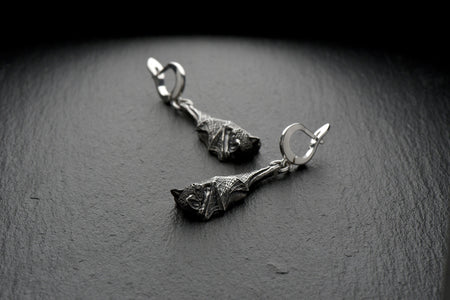 A high-resolution studio shot of a pair of dangle bat earrings resting on a dark slate surface. Each earring features a highly detailed, three-dimensional sleeping bat folded securely within its webbed wings. The 925 sterling silver is heavily oxidized in the recesses to highlight the intricate texture of the fur and wings. They connect to sturdy, polished silver lever-back clasps for secure wear.