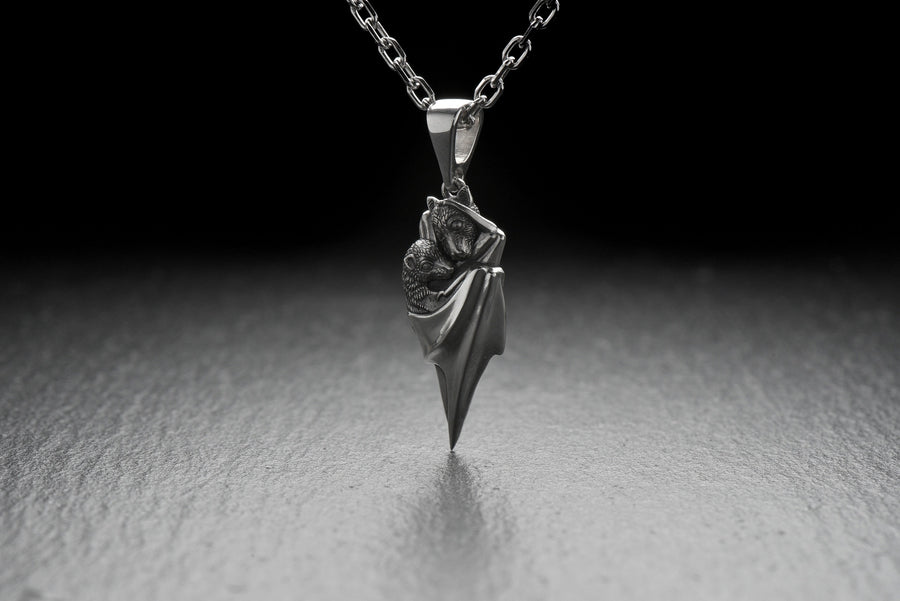 A detailed close-up studio shot of a sterling silver pendant shaped like two flying foxes or fruit bats. The bats are intimately huddled together, wrapped within the smooth, sweeping folds of their leathery wings. The metal is highly textured to show the fine details of their fur and faces. The silver is heavily oxidized in the recesses, creating a striking contrast against the polished highlights of the wings. (etsy image id: 5592925539)
