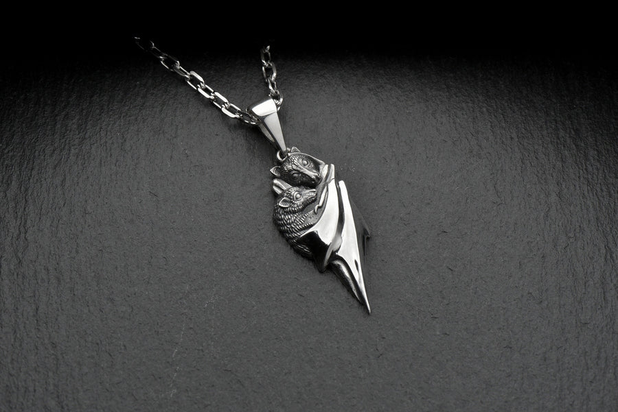 A detailed close-up studio shot of a sterling silver pendant shaped like two flying foxes or fruit bats. The bats are intimately huddled together, wrapped within the smooth, sweeping folds of their leathery wings. The metal is highly textured to show the fine details of their fur and faces. The silver is heavily oxidized in the recesses, creating a striking contrast against the polished highlights of the wings. (etsy image id: 5592925539)