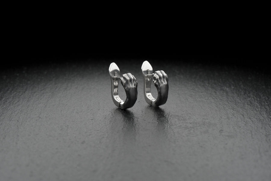 A close-up studio shot of a pair of sterling silver huggie hoop earrings resting on a dark slate surface. The front of each earring is masterfully sculpted to resemble a realistic cat paw, complete with distinct knuckles, claws, and soft paw pads. The earrings feature a hinged mechanism that allows the paw to wrap seamlessly around the earlobe. The metal is treated with heavy black oxidation in the recesses, contrasting beautifully with the brigh (etsy image id: 4850613175)