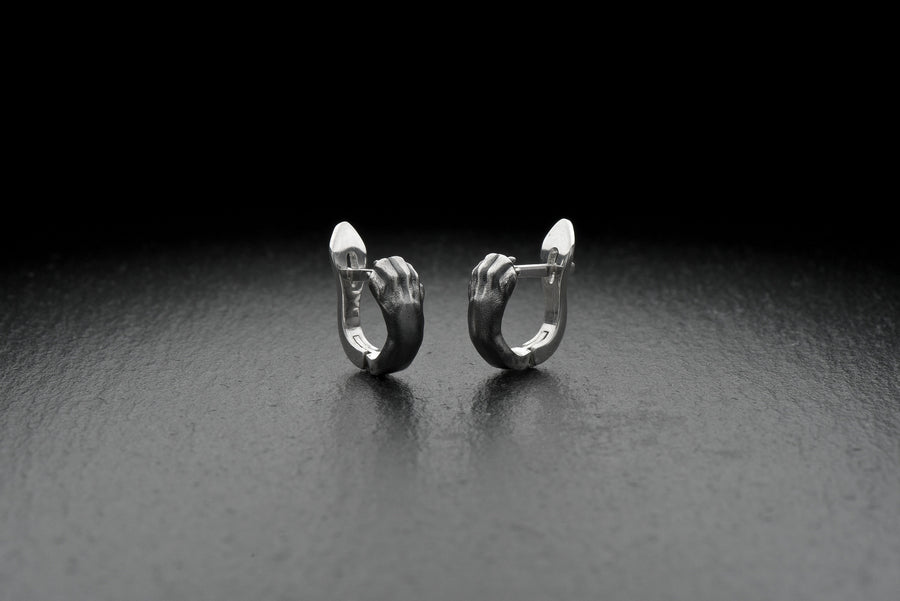 A close-up studio shot of a pair of sterling silver huggie hoop earrings resting on a dark slate surface. The front of each earring is masterfully sculpted to resemble a realistic cat paw, complete with distinct knuckles, claws, and soft paw pads. The earrings feature a hinged mechanism that allows the paw to wrap seamlessly around the earlobe. The metal is treated with heavy black oxidation in the recesses, contrasting beautifully with the brigh (etsy image id: 4850613175)