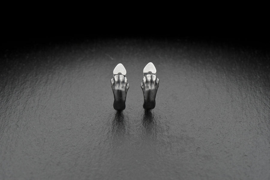 A close-up studio shot of a pair of sterling silver huggie hoop earrings resting on a dark slate surface. The front of each earring is masterfully sculpted to resemble a realistic cat paw, complete with distinct knuckles, claws, and soft paw pads. The earrings feature a hinged mechanism that allows the paw to wrap seamlessly around the earlobe. The metal is treated with heavy black oxidation in the recesses, contrasting beautifully with the brigh (etsy image id: 4850613175)