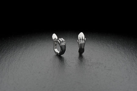 A close-up studio shot of a pair of sterling silver huggie hoop earrings resting on a dark slate surface. The front of each earring is masterfully sculpted to resemble a realistic cat paw, complete with distinct knuckles, claws, and soft paw pads. The earrings feature a hinged mechanism that allows the paw to wrap seamlessly around the earlobe. The metal is treated with heavy black oxidation in the recesses, contrasting beautifully with the brigh (etsy image id: 4850613175)