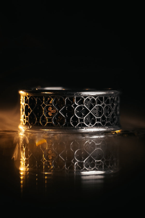 A high-definition shot of a matching two-piece wedding ring set resting on dark velvet. The foreground features a delicate sterling silver ring with a pointed, kite-shaped face holding a square blue gemstone. Standing behind it is a wide, sturdy band with a repeating quatrefoil "clover" pattern. Both rings share a cohesive finish: bright polished silver highlights against a deeply oxidized, matte black background.