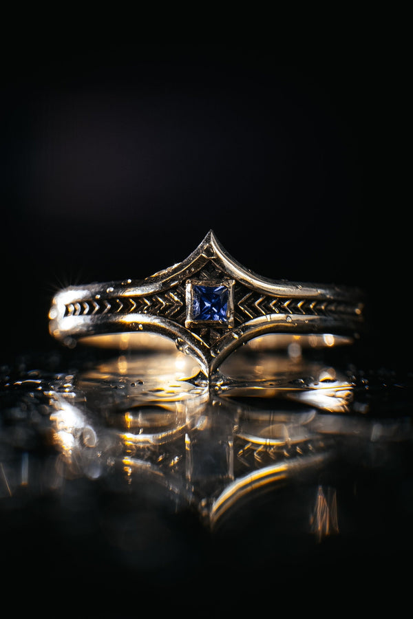 A high-definition shot of a matching two-piece wedding ring set resting on dark velvet. The foreground features a delicate sterling silver ring with a pointed, kite-shaped face holding a square blue gemstone. Standing behind it is a wide, sturdy band with a repeating quatrefoil "clover" pattern. Both rings share a cohesive finish: bright polished silver highlights against a deeply oxidized, matte black background.