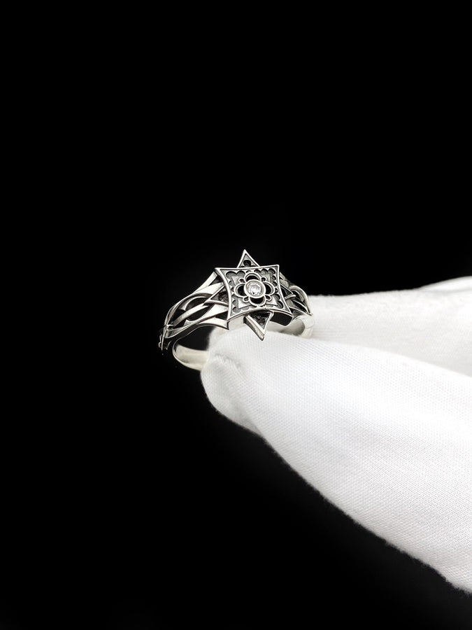 A detailed studio shot of a sterling silver ring held in a gloved hand. The focal point is a geometric "North Star" face—a square frame turned on its point, embellished with top and bottom spikes. Inside the frame is a floral quatrefoil pattern surrounding a round, bezel-set white gemstone (Diamond or CZ). The silver is polished on the raised lines and dark in the recesses. (etsy image id: 7452221522)