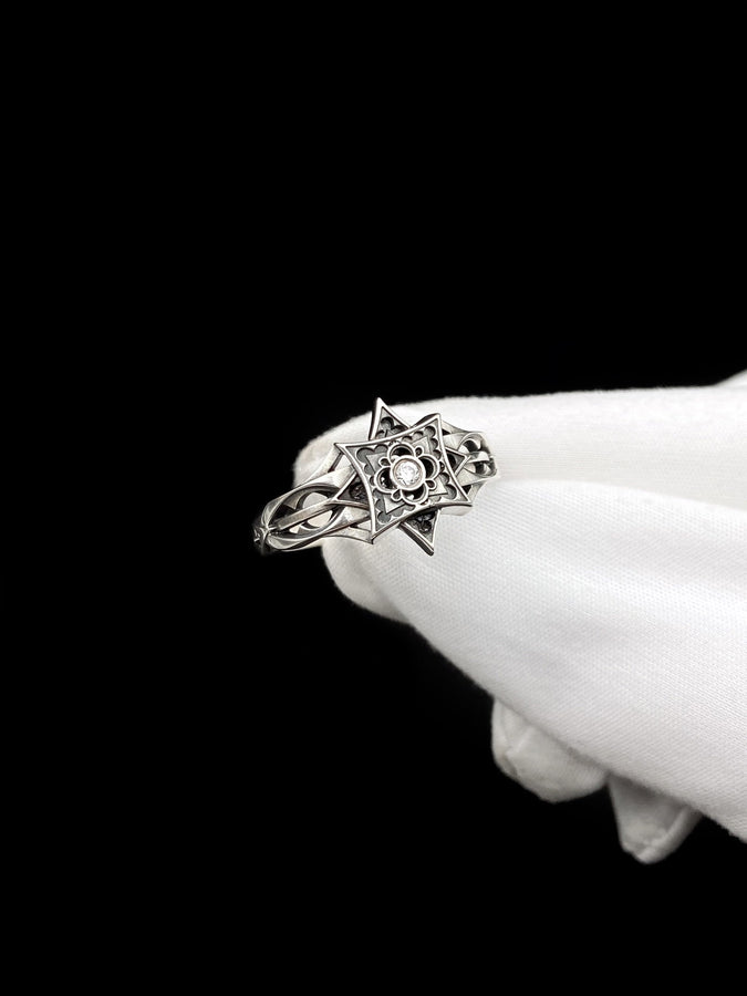 A detailed studio shot of a sterling silver ring held in a gloved hand. The focal point is a geometric "North Star" face—a square frame turned on its point, embellished with top and bottom spikes. Inside the frame is a floral quatrefoil pattern surrounding a round, bezel-set white gemstone (Diamond or CZ). The silver is polished on the raised lines and dark in the recesses. (etsy image id: 7452221522)
