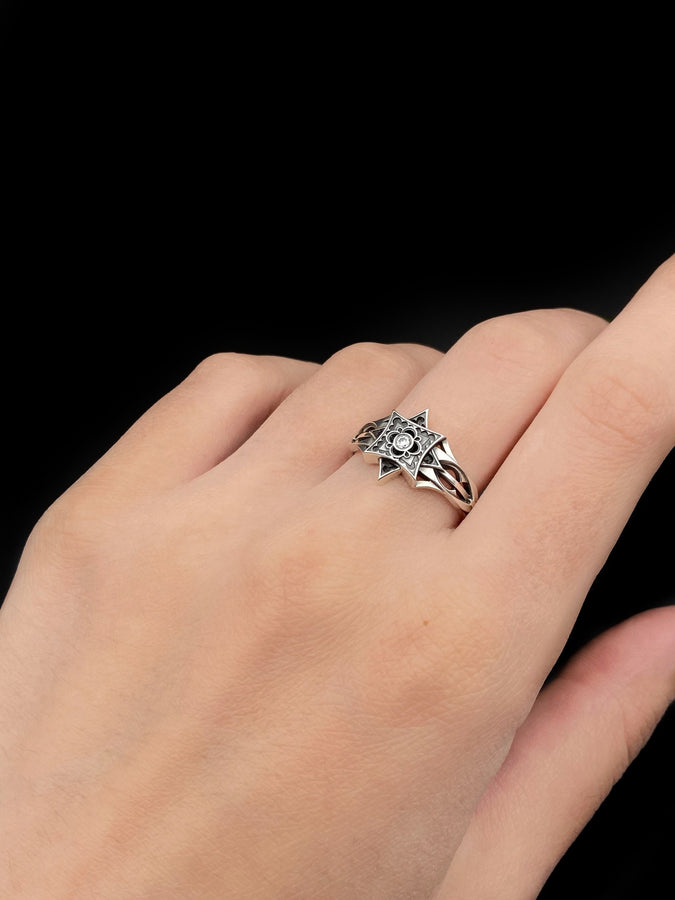 A detailed studio shot of a sterling silver ring held in a gloved hand. The focal point is a geometric "North Star" face—a square frame turned on its point, embellished with top and bottom spikes. Inside the frame is a floral quatrefoil pattern surrounding a round, bezel-set white gemstone (Diamond or CZ). The silver is polished on the raised lines and dark in the recesses. (etsy image id: 7452221522)