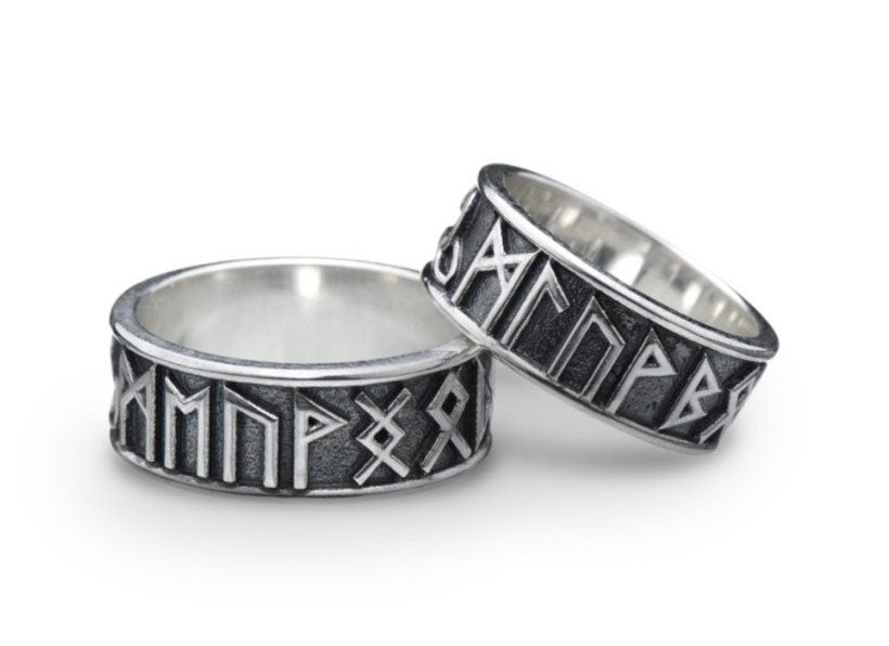 A clean studio shot of two matching sterling silver rings. The bands feature the 24 characters of the Elder Futhark runic alphabet wrapping around the exterior. The runes are polished silver, standing out sharply against a rough, blackened background texture. The rims are smooth and polished, framing the ancient script.