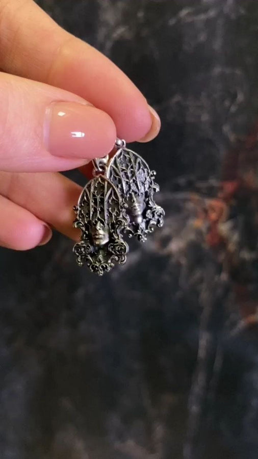 A detailed studio shot of a sterling silver pendant resting on a dark, textured stone surface. The design features a realistic human face in the center, but the forehead and eyes are obscured by a grid of Gothic window tracery (vertical bars and trefoils). The chin is framed by swirling Art Nouveau-style acanthus leaves. The silver is heavily oxidized, giving the face the appearance of a weathered stone statue. (etsy image id: 7448678222)