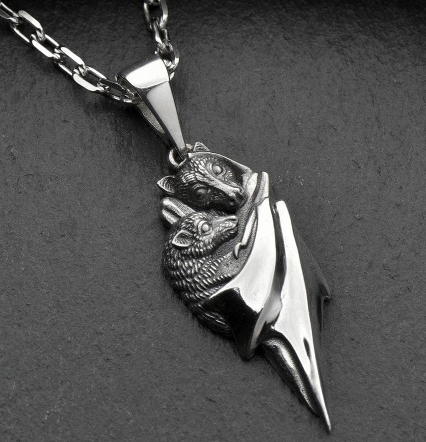A detailed close-up studio shot of a sterling silver pendant shaped like two flying foxes or fruit bats. The bats are intimately huddled together, wrapped within the smooth, sweeping folds of their leathery wings. The metal is highly textured to show the fine details of their fur and faces. The silver is heavily oxidized in the recesses, creating a striking contrast against the polished highlights of the wings. (etsy image id: 5592925539)