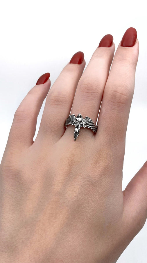 A clear studio shot of a sculptural sterling silver ring on a white background. The ring features a full-body female figure, likely an angel or goddess, positioned at the center. Her large, detailed wings spread horizontally to form the top half of the ring band. Her dress flows downward, tapering into the bottom of the band. (etsy image id: 7500519831)