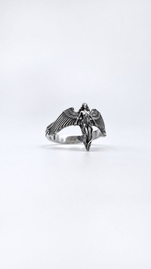A clear studio shot of a sculptural sterling silver ring on a white background. The ring features a full-body female figure, likely an angel or goddess, positioned at the center. Her large, detailed wings spread horizontally to form the top half of the ring band. Her dress flows downward, tapering into the bottom of the band. (etsy image id: 7500519831)