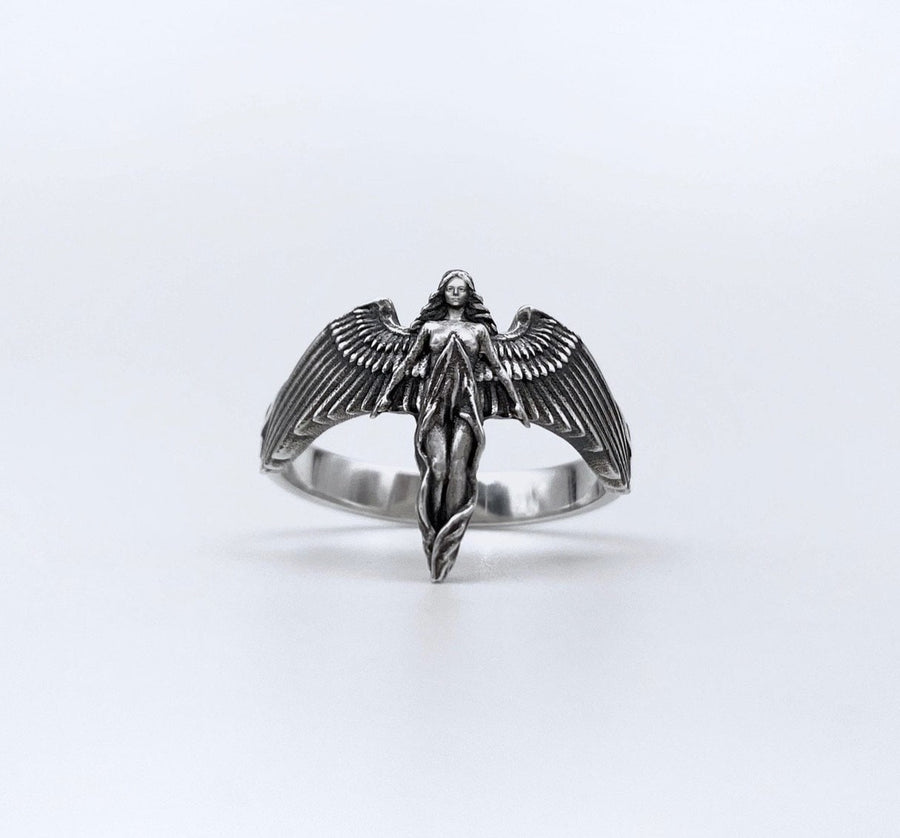 A clear studio shot of a sculptural sterling silver ring on a white background. The ring features a full-body female figure, likely an angel or goddess, positioned at the center. Her large, detailed wings spread horizontally to form the top half of the ring band. Her dress flows downward, tapering into the bottom of the band. (etsy image id: 7500519831)