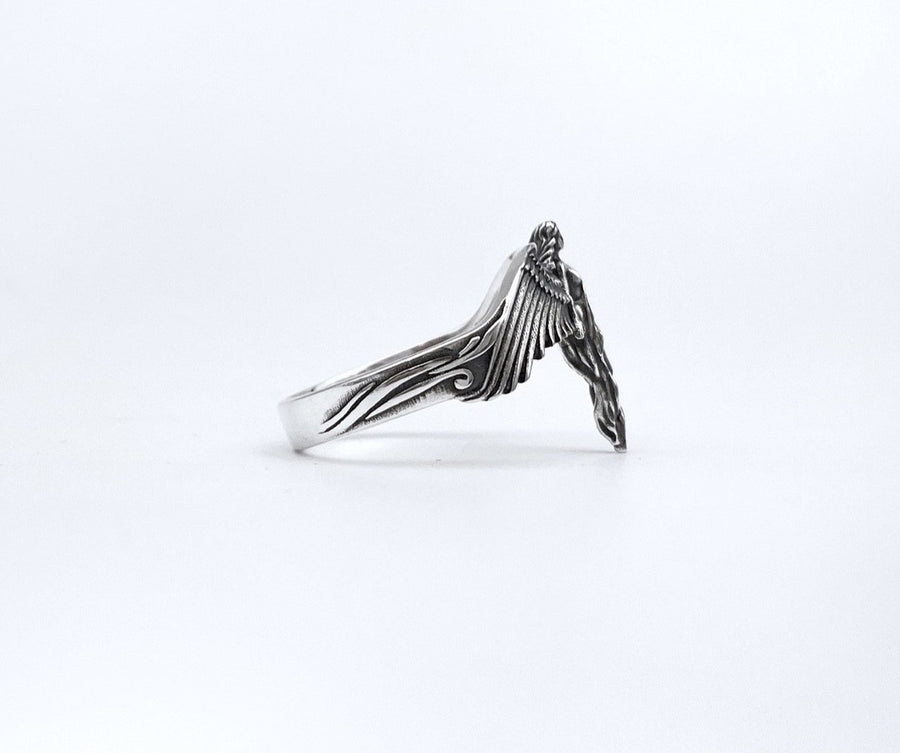 A clear studio shot of a sculptural sterling silver ring on a white background. The ring features a full-body female figure, likely an angel or goddess, positioned at the center. Her large, detailed wings spread horizontally to form the top half of the ring band. Her dress flows downward, tapering into the bottom of the band. (etsy image id: 7500519831)
