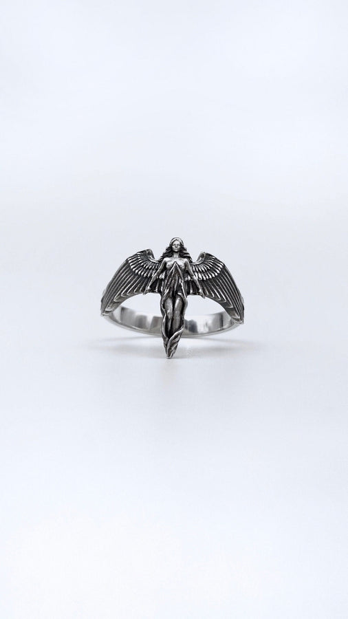 A clear studio shot of a sculptural sterling silver ring on a white background. The ring features a full-body female figure, likely an angel or goddess, positioned at the center. Her large, detailed wings spread horizontally to form the top half of the ring band. Her dress flows downward, tapering into the bottom of the band. (etsy image id: 7500519831)