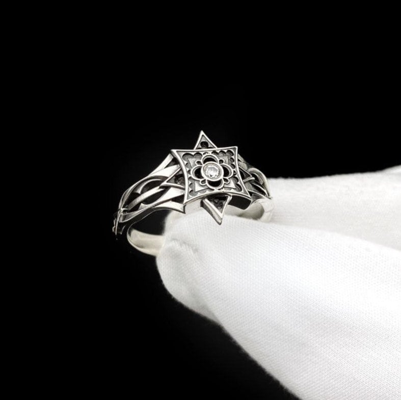 A detailed studio shot of a sterling silver ring held in a gloved hand. The focal point is a geometric "North Star" face—a square frame turned on its point, embellished with top and bottom spikes. Inside the frame is a floral quatrefoil pattern surrounding a round, bezel-set white gemstone (Diamond or CZ). The silver is polished on the raised lines and dark in the recesses. (etsy image id: 7452221522)