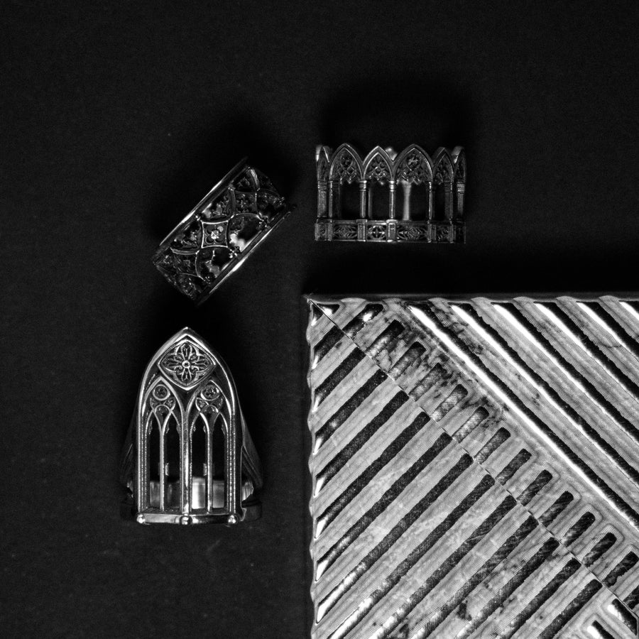 A clean studio shot of a tall, architectural sterling silver ring standing on a matte grey surface. The ring creates the silhouette of a Gothic "Lancet Window." It features three vertical openings separated by slender bars (mullions), topped by an intricate geometric tracery design within the pointed arch. The metal is matte silver with dark oxidation in the corners. (etsy image id: 7500328125)