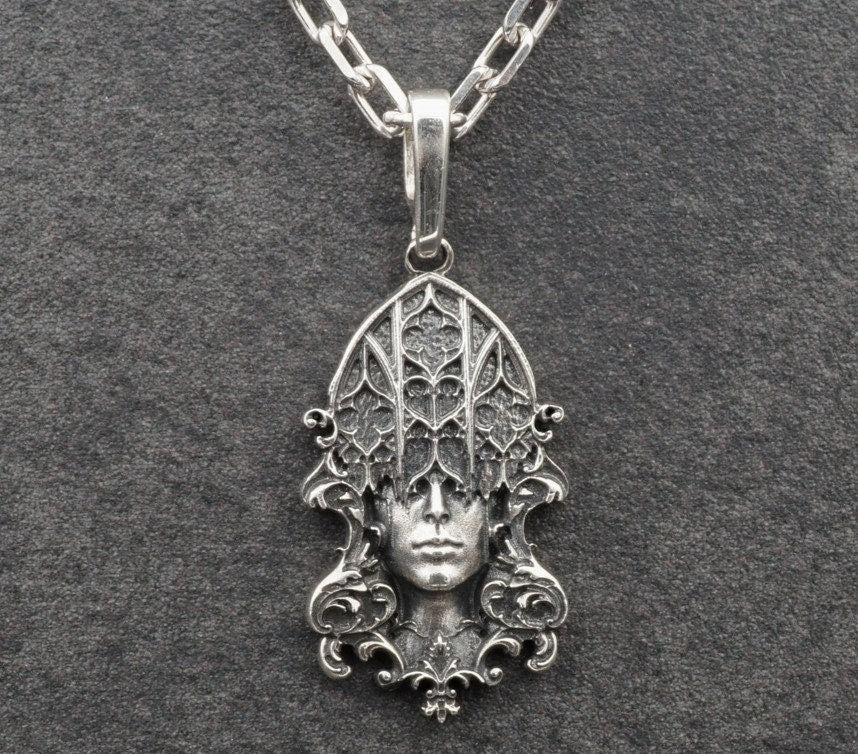 A detailed studio shot of a sterling silver pendant resting on a dark, textured stone surface. The design features a realistic human face in the center, but the forehead and eyes are obscured by a grid of Gothic window tracery (vertical bars and trefoils). The chin is framed by swirling Art Nouveau-style acanthus leaves. The silver is heavily oxidized, giving the face the appearance of a weathered stone statue. (etsy image id: 7448678222)