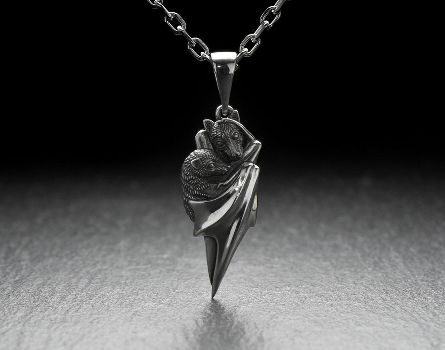 A detailed close-up studio shot of a sterling silver pendant shaped like two flying foxes or fruit bats. The bats are intimately huddled together, wrapped within the smooth, sweeping folds of their leathery wings. The metal is highly textured to show the fine details of their fur and faces. The silver is heavily oxidized in the recesses, creating a striking contrast against the polished highlights of the wings. (etsy image id: 5592925539)