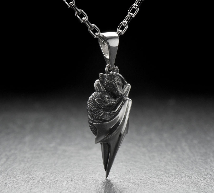 A detailed close-up studio shot of a sterling silver pendant shaped like two flying foxes or fruit bats. The bats are intimately huddled together, wrapped within the smooth, sweeping folds of their leathery wings. The metal is highly textured to show the fine details of their fur and faces. The silver is heavily oxidized in the recesses, creating a striking contrast against the polished highlights of the wings. (etsy image id: 5592925539)