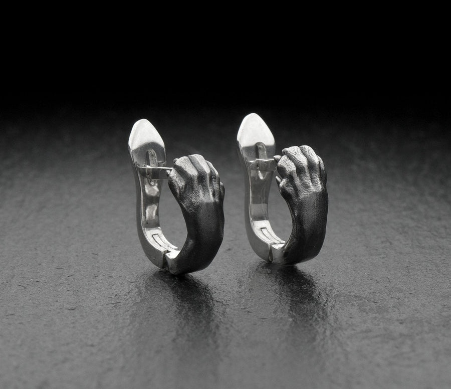 A close-up studio shot of a pair of sterling silver huggie hoop earrings resting on a dark slate surface. The front of each earring is masterfully sculpted to resemble a realistic cat paw, complete with distinct knuckles, claws, and soft paw pads. The earrings feature a hinged mechanism that allows the paw to wrap seamlessly around the earlobe. The metal is treated with heavy black oxidation in the recesses, contrasting beautifully with the brigh (etsy image id: 4850613175)