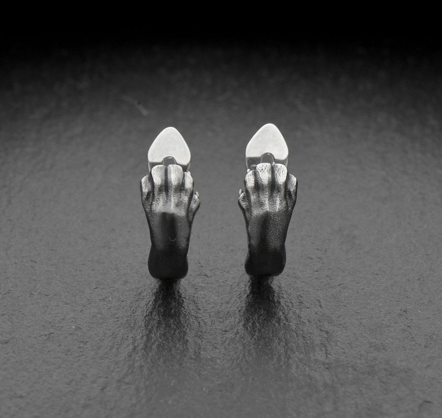 A close-up studio shot of a pair of sterling silver huggie hoop earrings resting on a dark slate surface. The front of each earring is masterfully sculpted to resemble a realistic cat paw, complete with distinct knuckles, claws, and soft paw pads. The earrings feature a hinged mechanism that allows the paw to wrap seamlessly around the earlobe. The metal is treated with heavy black oxidation in the recesses, contrasting beautifully with the brigh (etsy image id: 4850613175)