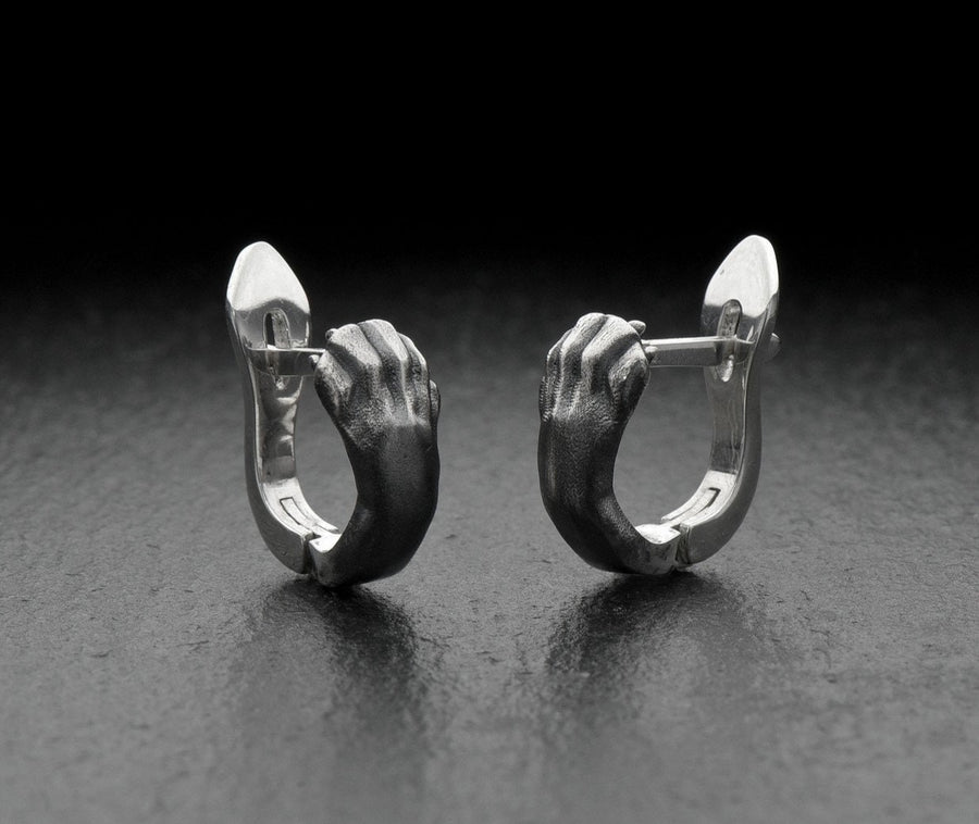 A close-up studio shot of a pair of sterling silver huggie hoop earrings resting on a dark slate surface. The front of each earring is masterfully sculpted to resemble a realistic cat paw, complete with distinct knuckles, claws, and soft paw pads. The earrings feature a hinged mechanism that allows the paw to wrap seamlessly around the earlobe. The metal is treated with heavy black oxidation in the recesses, contrasting beautifully with the brigh (etsy image id: 4850613175)