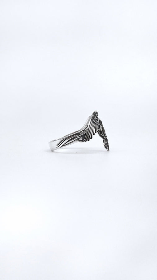 A clear studio shot of a sculptural sterling silver ring on a white background. The ring features a full-body female figure, likely an angel or goddess, positioned at the center. Her large, detailed wings spread horizontally to form the top half of the ring band. Her dress flows downward, tapering into the bottom of the band. (etsy image id: 7500519831)