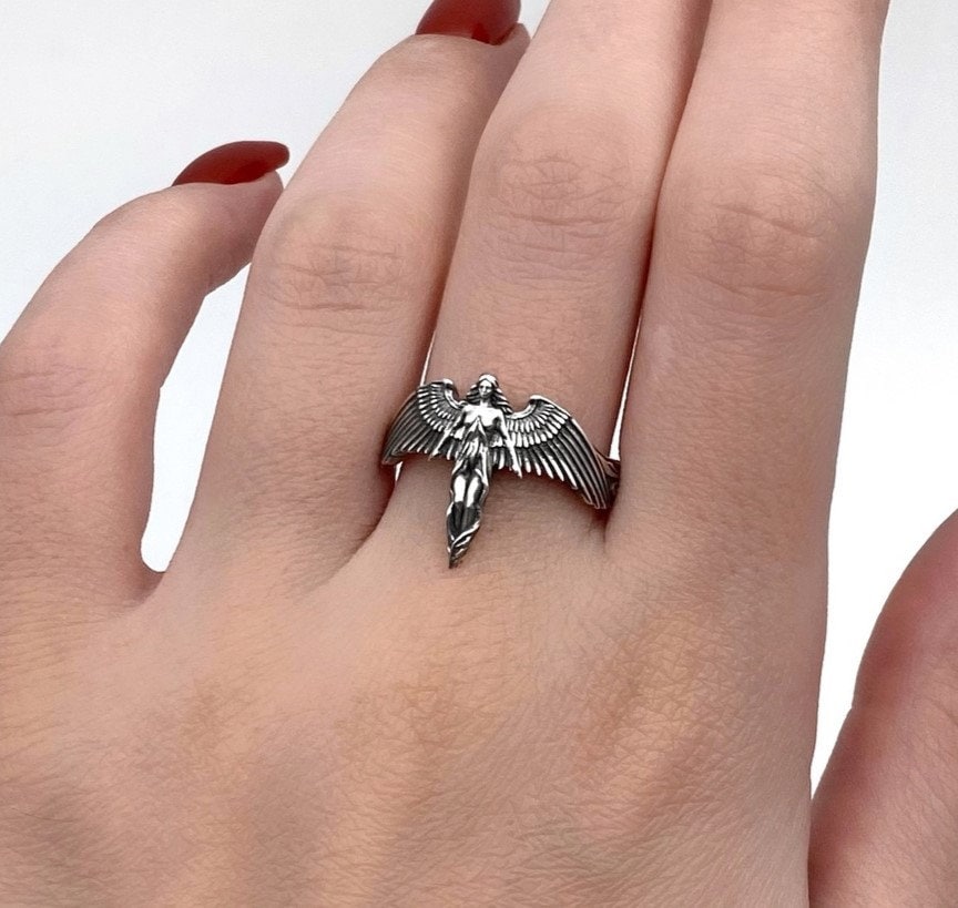 A clear studio shot of a sculptural sterling silver ring on a white background. The ring features a full-body female figure, likely an angel or goddess, positioned at the center. Her large, detailed wings spread horizontally to form the top half of the ring band. Her dress flows downward, tapering into the bottom of the band. (etsy image id: 7500519831)