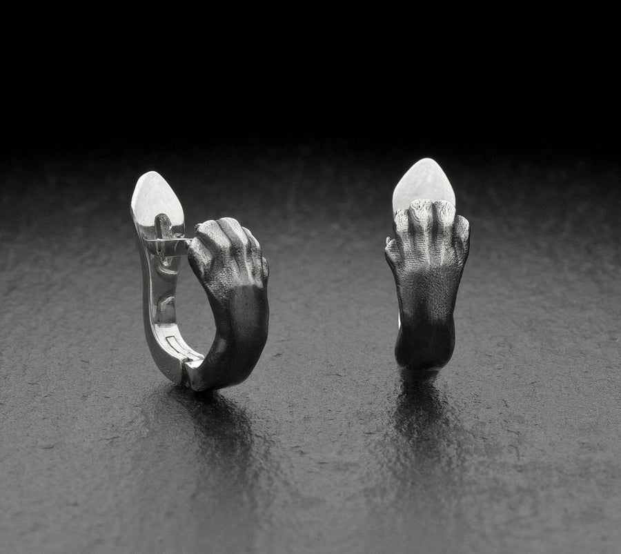 A close-up studio shot of a pair of sterling silver huggie hoop earrings resting on a dark slate surface. The front of each earring is masterfully sculpted to resemble a realistic cat paw, complete with distinct knuckles, claws, and soft paw pads. The earrings feature a hinged mechanism that allows the paw to wrap seamlessly around the earlobe. The metal is treated with heavy black oxidation in the recesses, contrasting beautifully with the brigh (etsy image id: 4850613175)