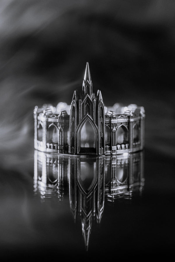 A cinematic studio shot of a sculptural sterling silver ring standing on a reflective surface. The ring is shaped like a miniature building, featuring a tall central spire that rises to a sharp point. The band itself is formed by a colonnade of pointed Gothic arches and buttresses that wrap around the finger. The silver is heavily oxidized, creating deep shadows in the "windows" and "doorways" of the structure. (etsy image id: 7450125356)
