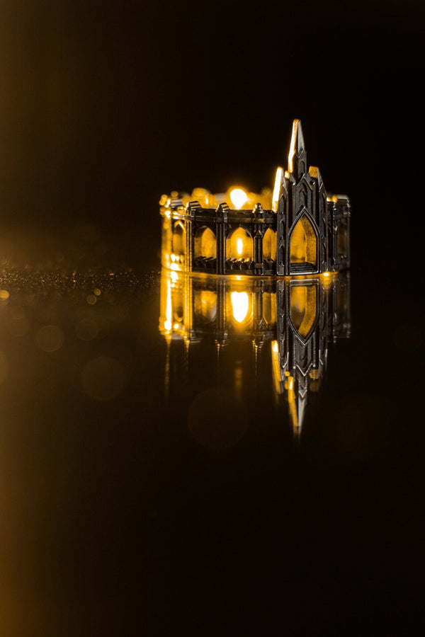 A cinematic studio shot of a sculptural sterling silver ring standing on a reflective surface. The ring is shaped like a miniature building, featuring a tall central spire that rises to a sharp point. The band itself is formed by a colonnade of pointed Gothic arches and buttresses that wrap around the finger. The silver is heavily oxidized, creating deep shadows in the "windows" and "doorways" of the structure. (etsy image id: 7450125356)