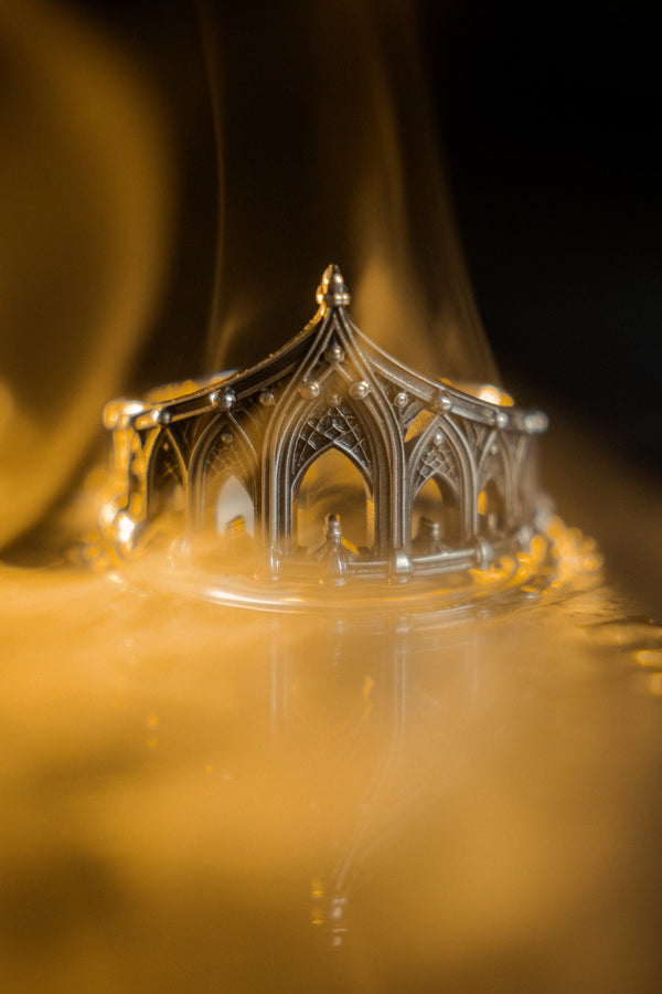 A cinematic studio shot of a sculptural sterling silver ring standing on a reflective surface. The ring is designed as a "crown" or "coronet," formed by a continuous circle of Gothic cathedral arches. The central arch is the tallest, creating a focal point like a spire. The silver is heavily oxidized, with black recesses emphasizing the ribbed vaulting and architectural details against the polished ridges.