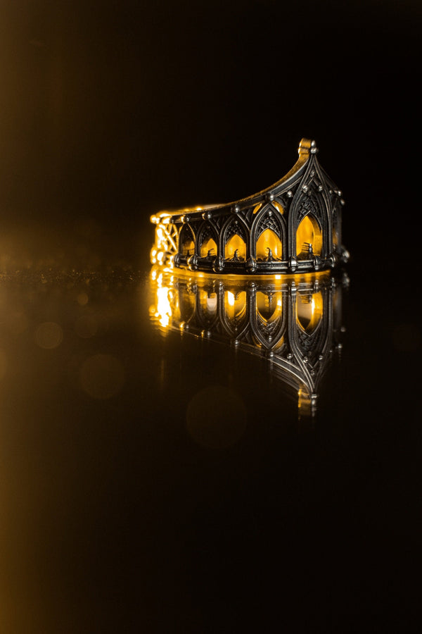 A cinematic studio shot of a sculptural sterling silver ring standing on a reflective surface. The ring is designed as a "crown" or "coronet," formed by a continuous circle of Gothic cathedral arches. The central arch is the tallest, creating a focal point like a spire. The silver is heavily oxidized, with black recesses emphasizing the ribbed vaulting and architectural details against the polished ridges.