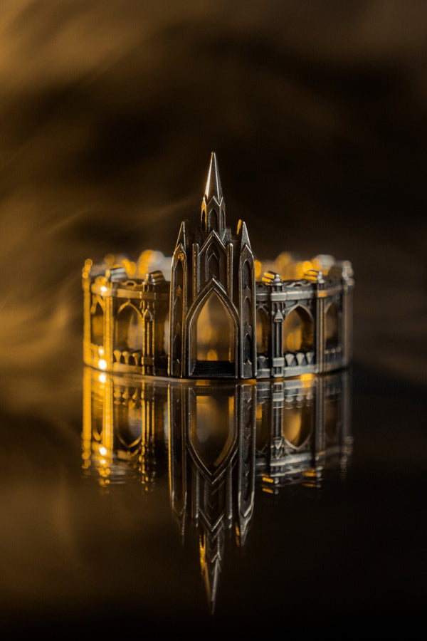 A cinematic studio shot of a sculptural sterling silver ring standing on a reflective surface. The ring is shaped like a miniature building, featuring a tall central spire that rises to a sharp point. The band itself is formed by a colonnade of pointed Gothic arches and buttresses that wrap around the finger. The silver is heavily oxidized, creating deep shadows in the "windows" and "doorways" of the structure. (etsy image id: 7450125356)