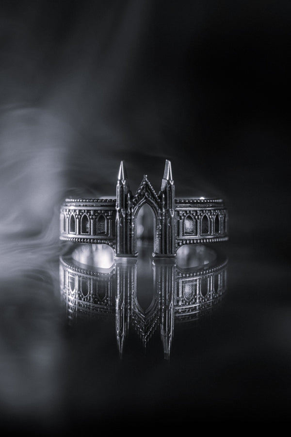A cinematic studio shot of a sculptural sterling silver ring standing on a reflective surface. The design mimics the facade of a Gothic cathedral or castle gate. Two tall, pointed spires rise up on either side of a central archway. The band continues outwards with a colonnade of smaller arches. The silver features a beaded "cornice" detail along the edges and is heavily oxidized to emphasize the depth of the "stonework." (etsy image id: 7450300962)