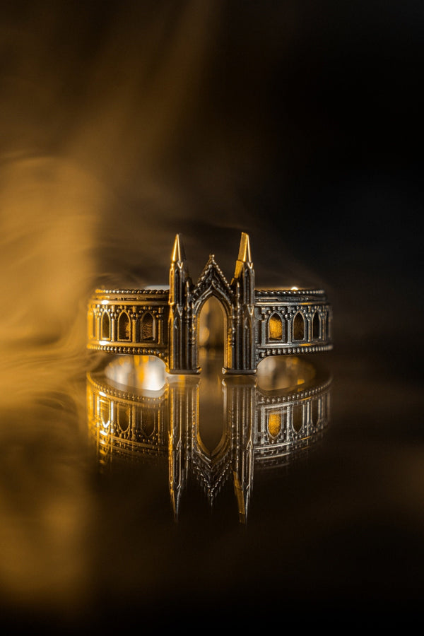 A cinematic studio shot of a sculptural sterling silver ring standing on a reflective surface. The design mimics the facade of a Gothic cathedral or castle gate. Two tall, pointed spires rise up on either side of a central archway. The band continues outwards with a colonnade of smaller arches. The silver features a beaded "cornice" detail along the edges and is heavily oxidized to emphasize the depth of the "stonework." (etsy image id: 7450300962)
