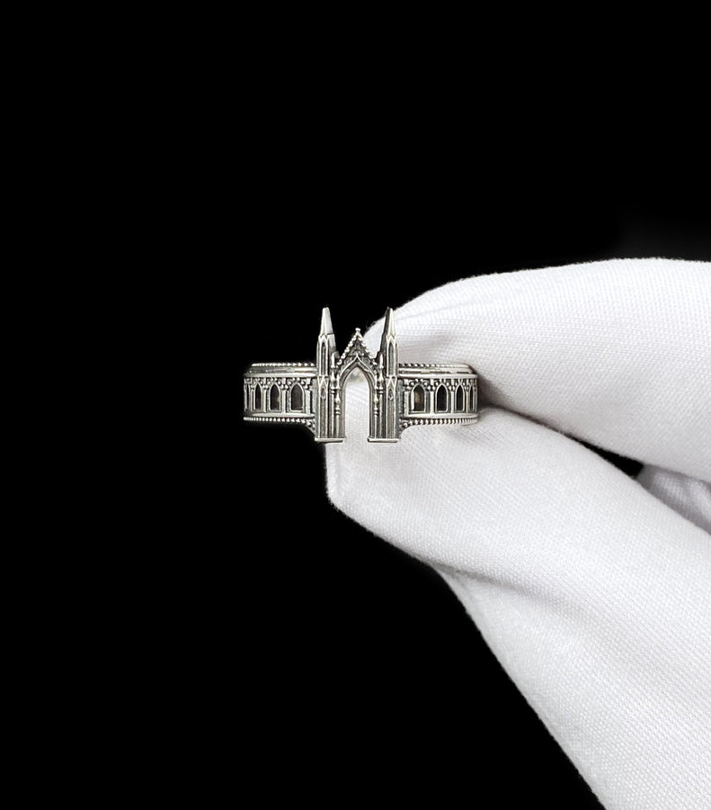 A cinematic studio shot of a sculptural sterling silver ring standing on a reflective surface. The design mimics the facade of a Gothic cathedral or castle gate. Two tall, pointed spires rise up on either side of a central archway. The band continues outwards with a colonnade of smaller arches. The silver features a beaded "cornice" detail along the edges and is heavily oxidized to emphasize the depth of the "stonework." (etsy image id: 7450300962)