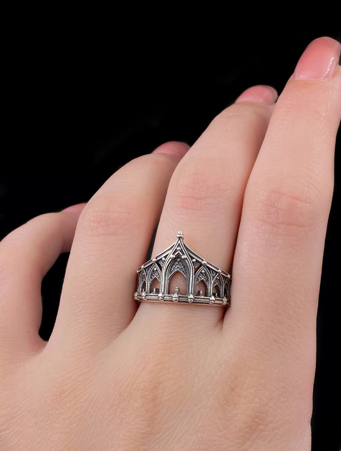 A cinematic studio shot of a sculptural sterling silver ring standing on a reflective surface. The ring is designed as a "crown" or "coronet," formed by a continuous circle of Gothic cathedral arches. The central arch is the tallest, creating a focal point like a spire. The silver is heavily oxidized, with black recesses emphasizing the ribbed vaulting and architectural details against the polished ridges.